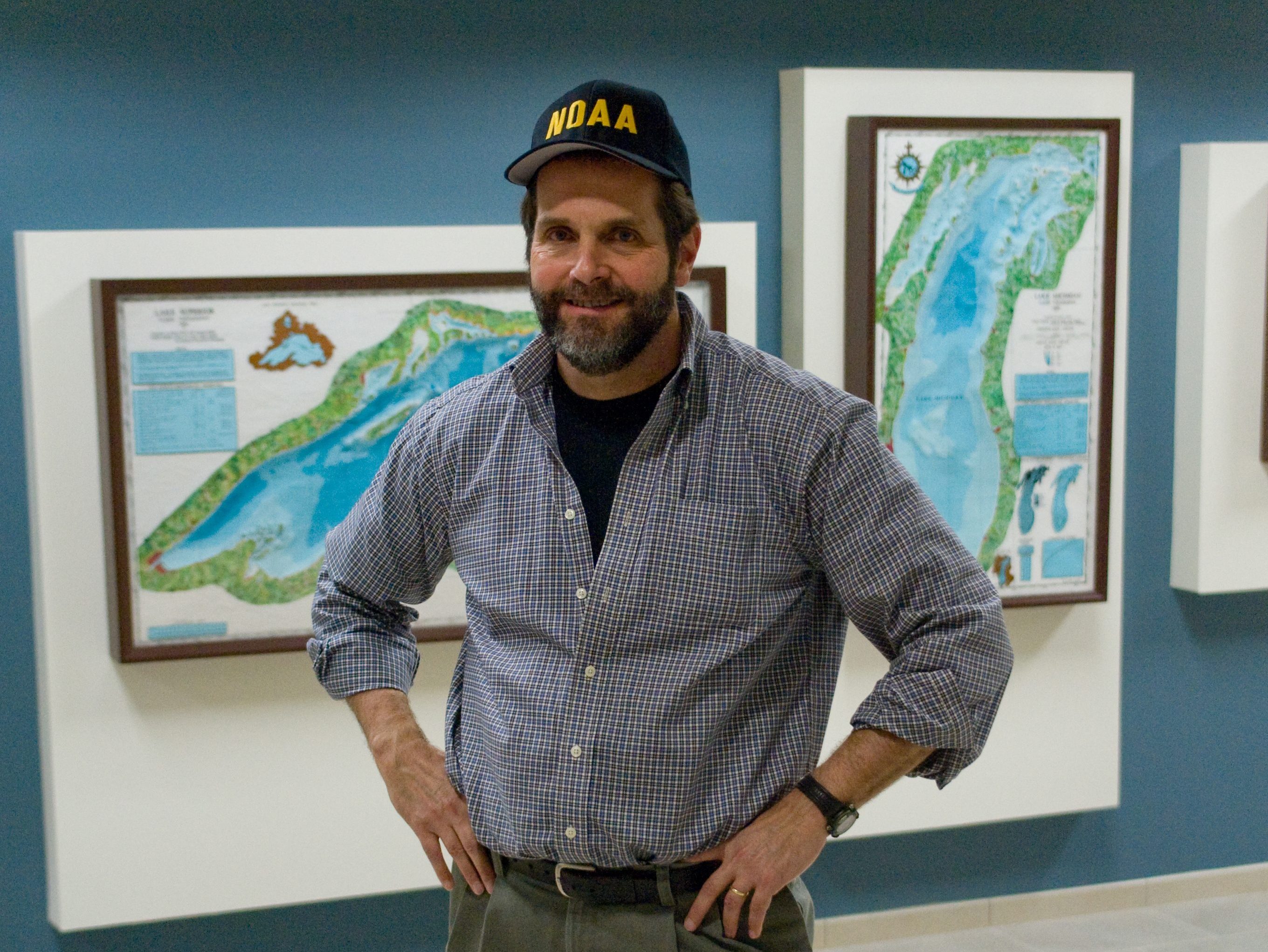 A man in a baseball cap stands in the GLERL lobby in front of some 3-d bathymetry maps of the Great Lakes