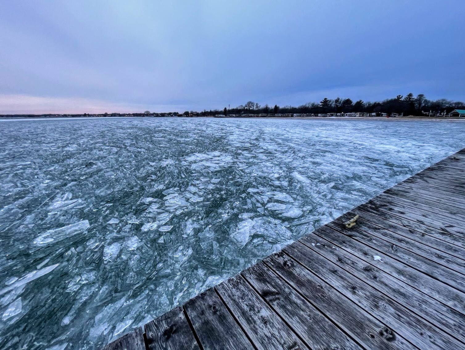 View looking out onto open water of lake Huron with ice chunks swirling in the lake. A wooden dock is seen in the foreground and the shoreline with trees and houses is in the distance.